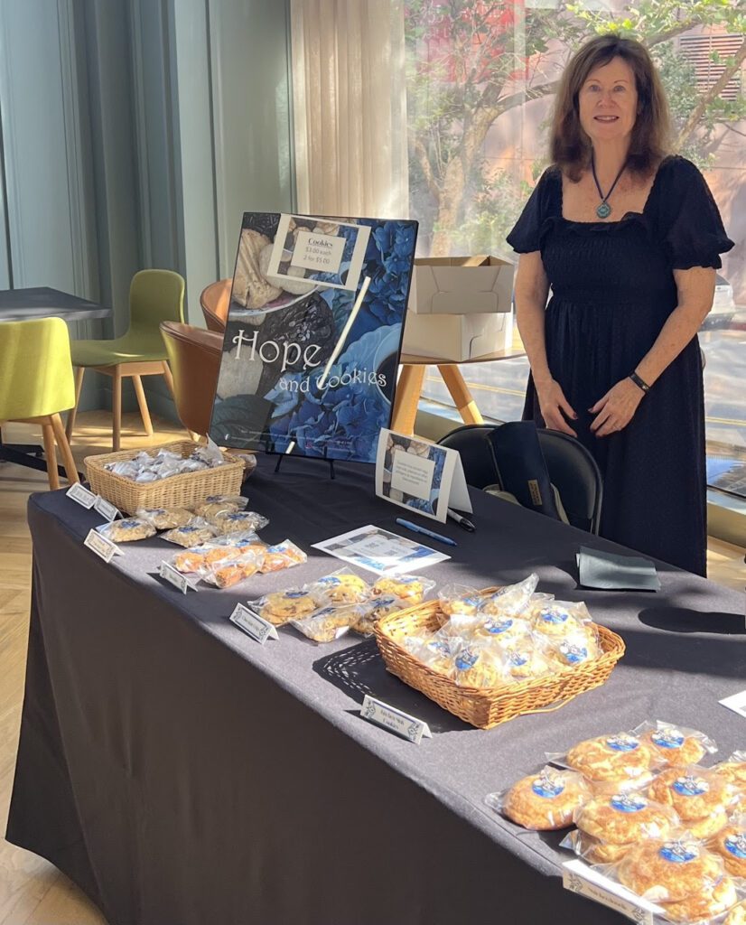 A woman in a black dress stands behind a table covered with baskets of cookies for sale. A sign reads 