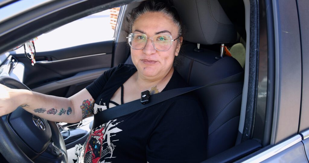 A person with glasses and tattoos on their arm sits in the driver's seat of a car, smiling at the camera. Wearing a black shirt and seatbelt, they appear ready to help with food distributions in their community.