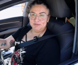 A person with glasses and tattoos on their arm sits in the driver's seat of a car, smiling at the camera. Wearing a black shirt and seatbelt, they appear ready to help with food distributions in their community.
