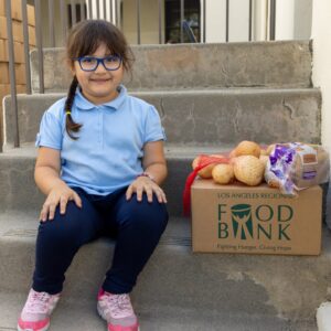 A young person sits on outdoor steps next to a cardboard box labeled "Los Angeles Regional Food Bank," containing groceries like onions and bread. She is smiling, wearing glasses, a light blue shirt, dark pants, and pink sneakers.