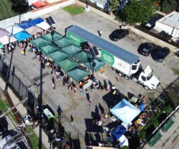 Aerial view of a Food Bank event with green and blue tents, a large white truck, and crowds gathered on a paved lot surrounded by fences and parked cars.