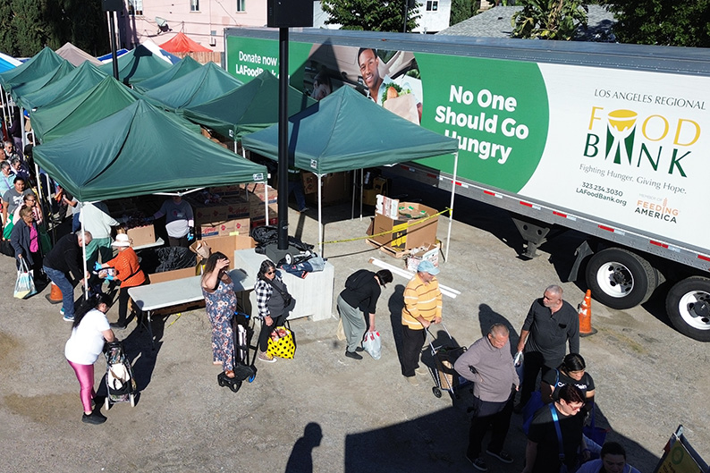 People wait in line at an outdoor WeFeedLA food distribution event next to a truck labeled "Los Angeles Regional Food Bank," with a banner reading "No One Should Go Hungry" as volunteers work under green tents.