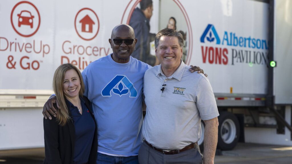 Three people stand smiling in front of a truck with Albertsons, Vons, and Pavilions logos, promoting DriveUp & Go, Grocery Delivery, and hunger relief services. The two men and one woman have their arms around each other.