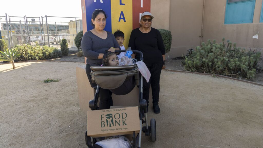 Two women and two children stand outside a building, one woman pushing a stroller with a child and a large Los Angeles Regional Food Bank box, highlighting food assistance. A colorful mural and plants decorate the background.