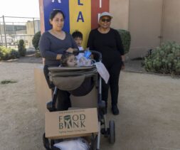 Two women and two children stand outside a building, one woman pushing a stroller with a child and a large Los Angeles Regional Food Bank box, highlighting food assistance. A colorful mural and plants decorate the background.