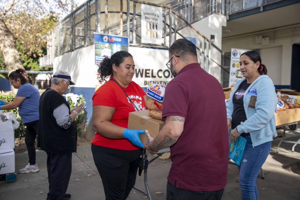 People are distributing and receiving food assistance outdoors. A woman in a red shirt hands a box and a loaf of bread to a man, while others sort and collect food at tables nearby. A 