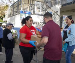 People are distributing and receiving food assistance outdoors. A woman in a red shirt hands a box and a loaf of bread to a man, while others sort and collect food at tables nearby. A 