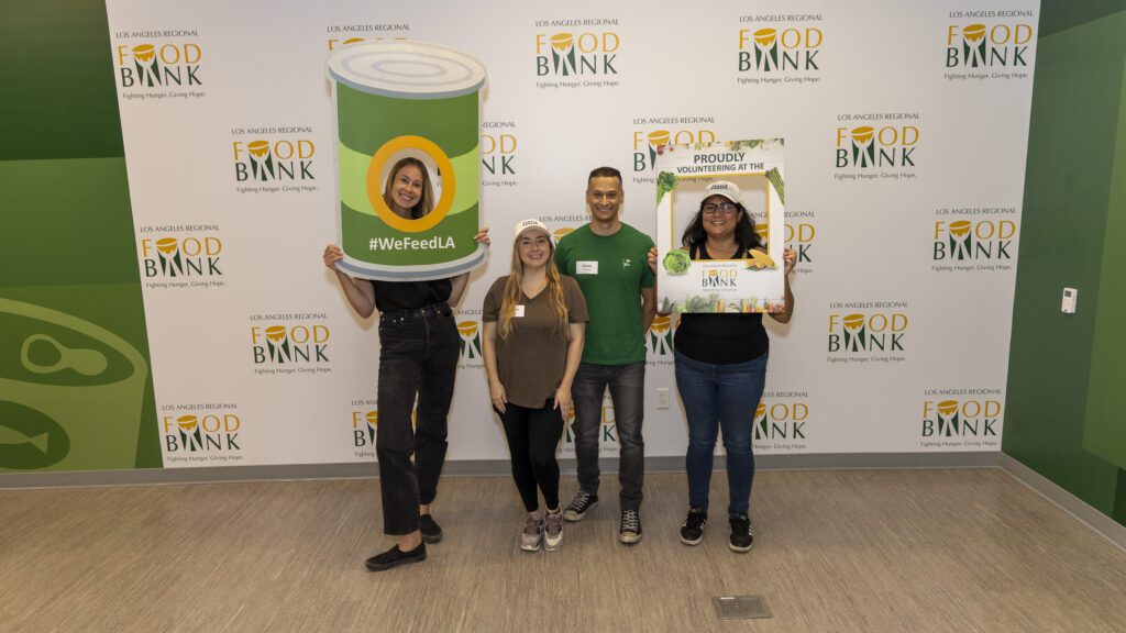 Four smiling volunteers pose at the Los Angeles Regional Food Bank, holding fun props including a giant can frame and a sign that says 