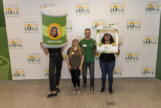 Four smiling volunteers pose at the Los Angeles Regional Food Bank, holding fun props including a giant can frame and a sign that says "PROUDLY VOLUNTEERING." The backdrop features the food bank’s logo.