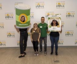 Four smiling volunteers pose at the Los Angeles Regional Food Bank, holding fun props including a giant can frame and a sign that says 