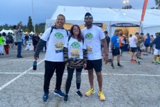 Three people wearing matching “Team WeFeedLA” race shirts and bibs stand smiling in a parking lot at a race event, with a large white tent and other participants in the background.