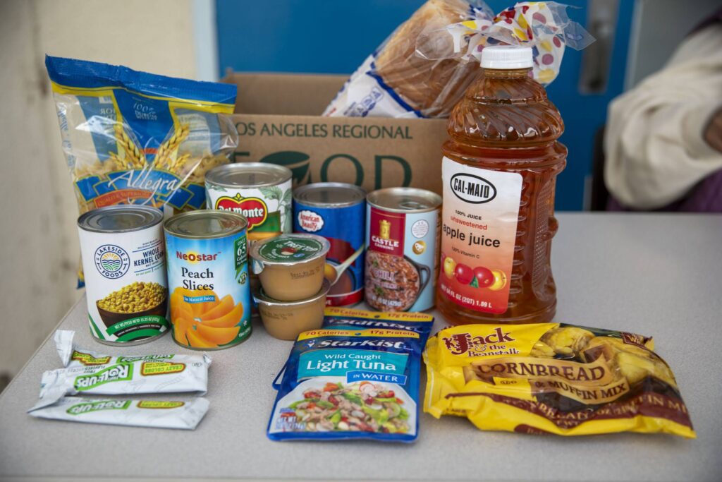 A variety of food items are arranged on a table, including canned vegetables and fruit, apple juice, bread, cornbread mix, tuna, applesauce cups, and granola bars—ready for donation to the local food bank. A food donation box sits in the background.