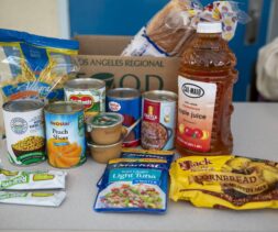 A variety of food items are arranged on a table, including canned vegetables and fruit, apple juice, bread, cornbread mix, tuna, applesauce cups, and granola bars—ready for donation to the local food bank. A food donation box sits in the background.