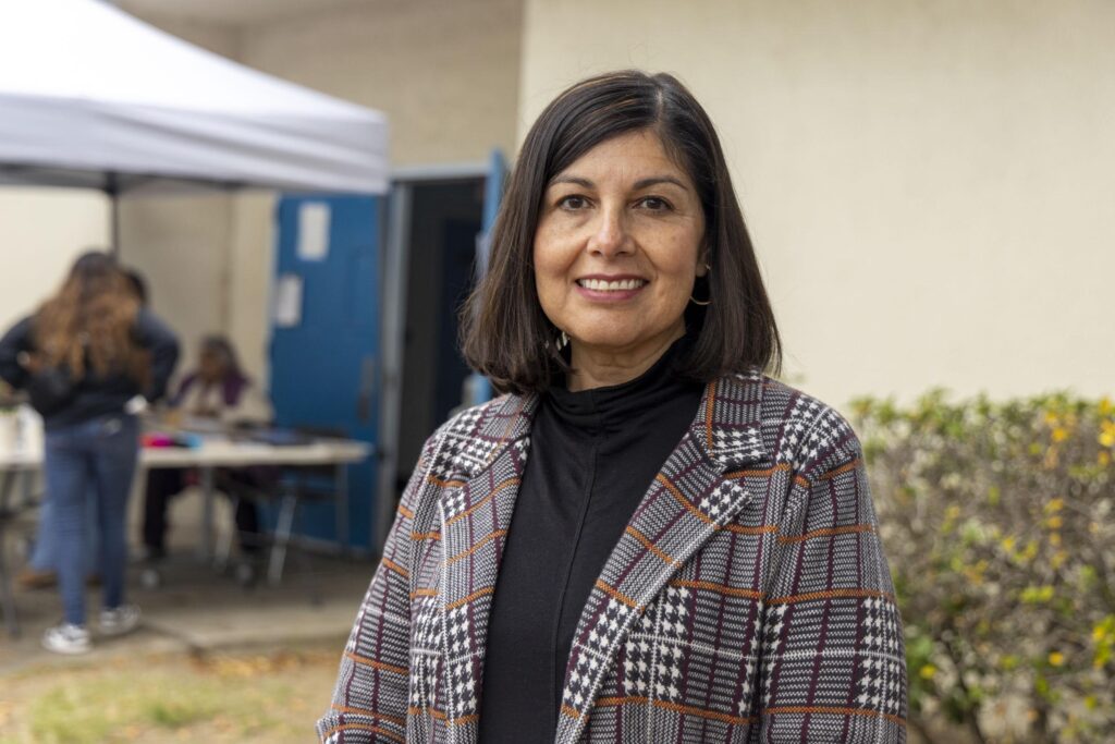 A woman with shoulder-length dark hair, wearing a plaid coat and black turtleneck, stands outdoors and smiles. In the background, people sit at a table under a canopy near a building, possibly attending a food bank event.