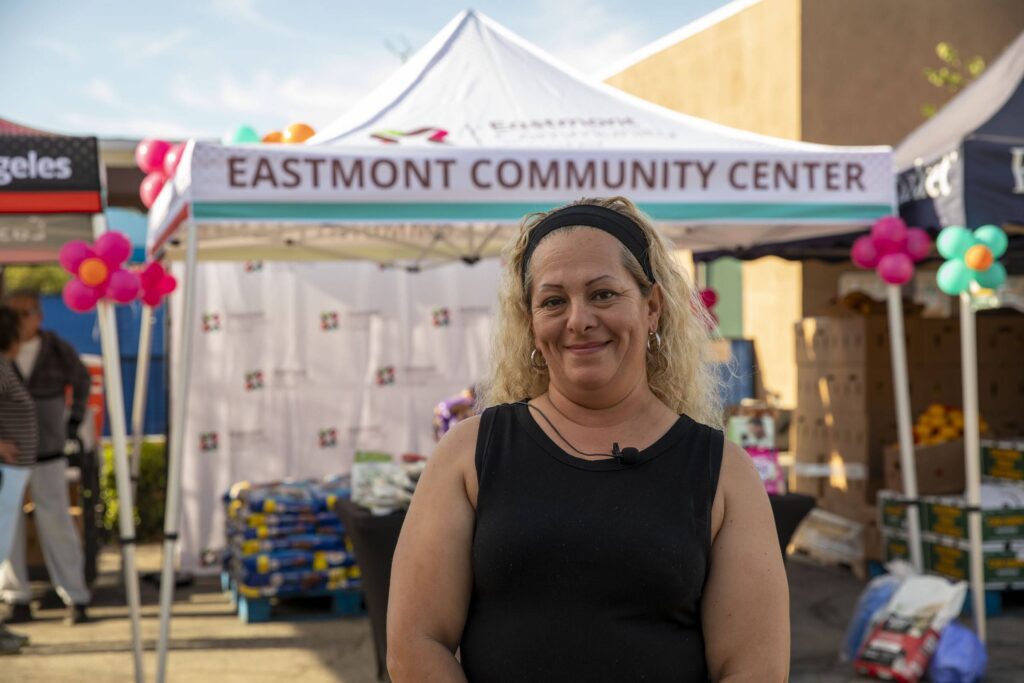 A woman with blonde hair and a headband smiles in front of the Eastmont Community Center tent at an outdoor community event, where boxes and supplies for the backpack program are visible behind her.