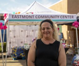 A woman with blonde hair and a headband smiles in front of the Eastmont Community Center tent at an outdoor community event, where boxes and supplies for the backpack program are visible behind her.