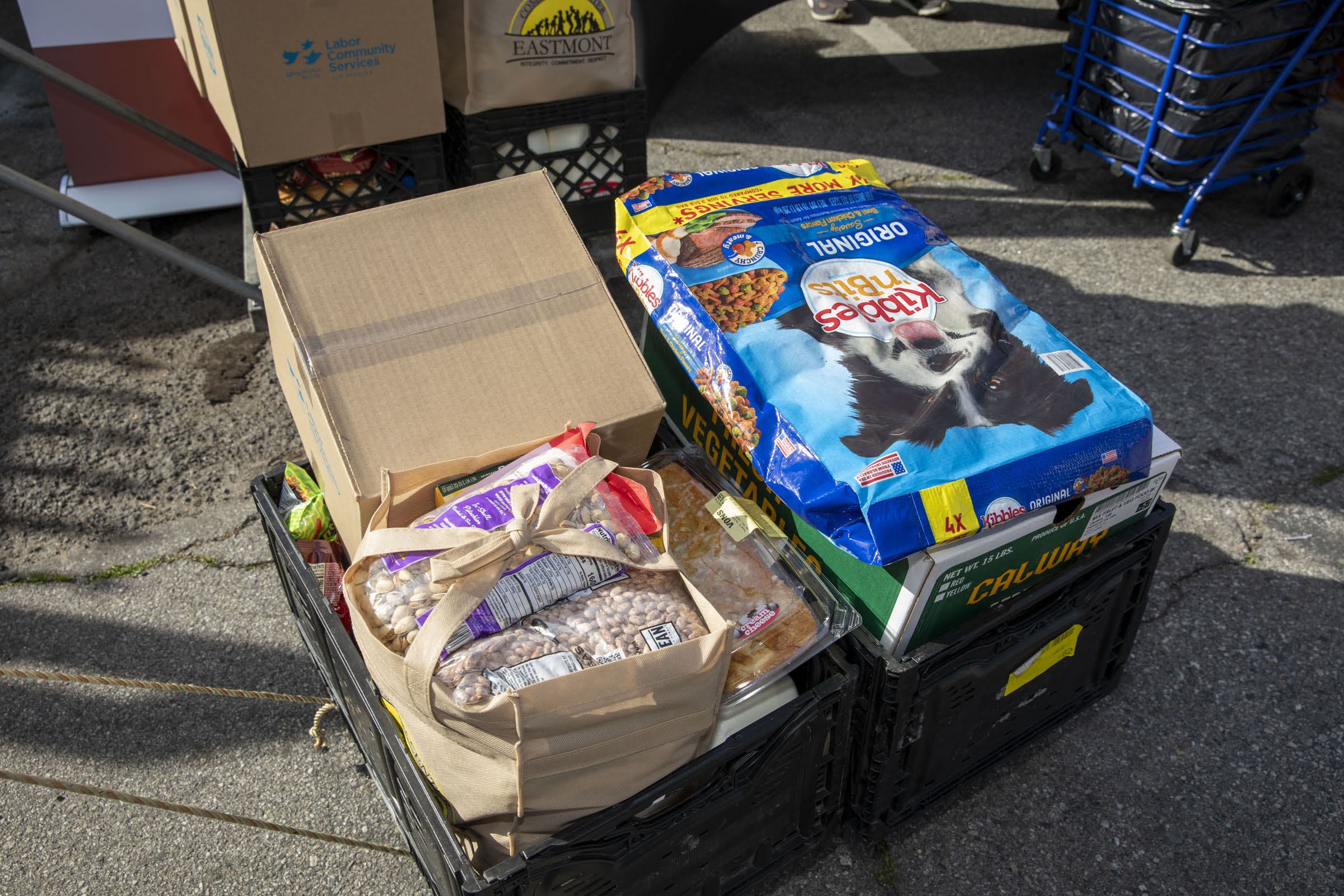 A crate on the ground holds a paper bag of groceries and a large bag of dog food, highlighting the impact of Assembly Bill 660. There are more boxes and crates, a blue shopping cart, and people in the background.
