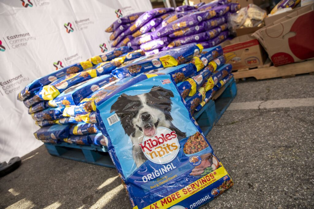 Bags of Kibbles 'n Bits Original dog food, in line with Assembly Bill 660 standards, are stacked on a blue pallet outdoors. One bag stands upright in front; other pet food bags and a white Eastmont Community Center banner appear behind.