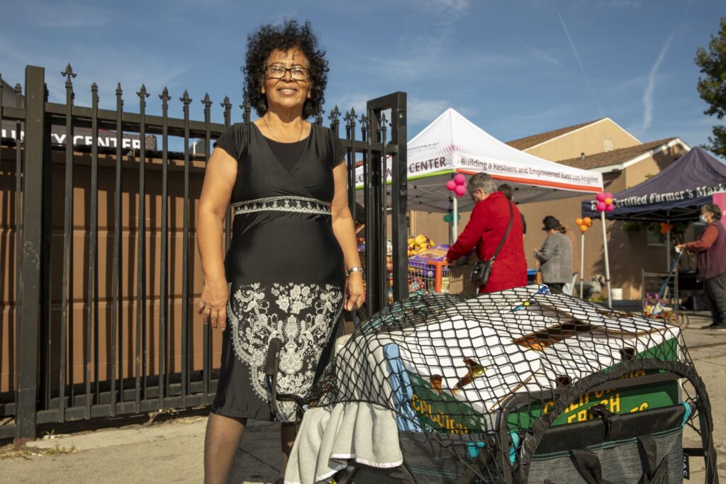 A smiling woman in a black and white dress stands outdoors by a fenced gate, next to a cart filled with bags and towels, possibly for local food pantries. Behind her, people gather near white tents at a sunny outdoor market or community event.