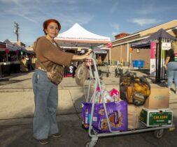 A woman with red hair stands outdoors holding a shopping cart filled with groceries, including oranges, cat food, and produce, at a street market where food distribution takes place amid tents and bustling vendors.