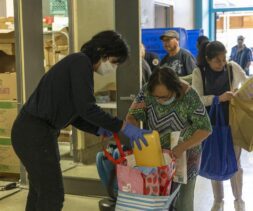 A volunteer wearing gloves and a mask helps a woman pack groceries and a food box into bags at a community food distribution event, while others wait in line holding bags.
