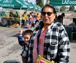A woman in sunglasses and a plaid jacket stands outdoors with a child beside her at a mobile food pantry event, with LA Food Bank tents and volunteers visible in the background.