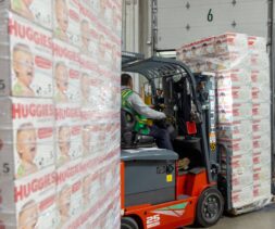A worker operates a forklift to move pallets stacked with boxes of Huggies diapers, part of a diaper assistance program, inside a warehouse. The boxes feature images of smiling babies.