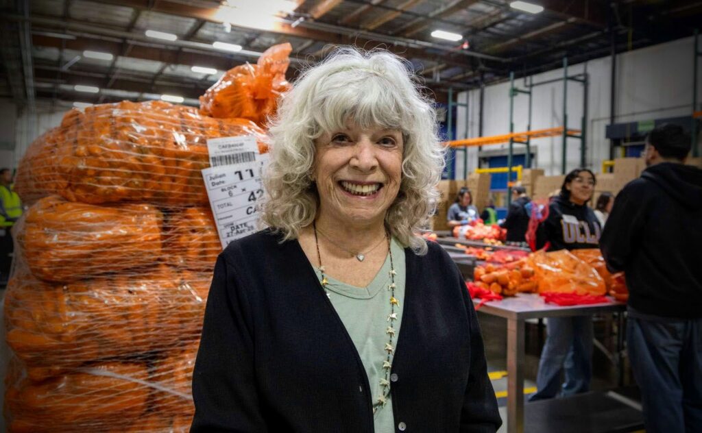 An older woman with gray curly hair smiles in a warehouse, standing before large bags of oranges. In the background, people sort produce on tables—part of a volunteer program that also offers tax benefits to participants.