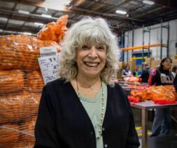An older woman with gray curly hair smiles in a warehouse, standing before large bags of oranges. In the background, people sort produce on tables—part of a volunteer program that also offers tax benefits to participants.