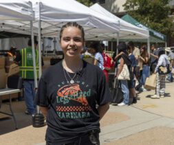 A person with braided hair smiles at the camera while standing outside near tents at a mobile food pantry. Several people are gathered in the background under the tents, some wearing backpacks and hats. It appears to be a sunny day.
