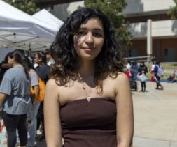 A young woman with wavy dark hair and a strapless brown top stands outdoors, smiling slightly. She has a purple marking on her left cheek. People, tents, and a mobile food pantry are visible in the background under sunny skies.