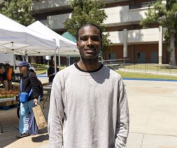 A man in a light gray long-sleeve shirt stands outdoors, smiling at the camera. Behind him, a mobile food pantry serves people under white tents, with tables of produce and a multi-story building framed by trees.