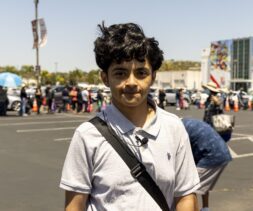 A young person with dark curly hair, wearing a light grey polo shirt and a black shoulder strap, stands in a sunny parking lot with a long line of people waiting at a mobile food pantry near a building in the background.