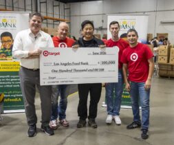 Five people, including a volunteer, smiling and holding a large $100,000 check made out to the Los Angeles Food Bank from Target. They stand in a warehouse with food bank banners and boxes in the background.
