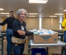 An older man with gray hair smiles while sorting packaged goods into a box labeled 