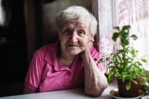 An older woman wearing pink sits next to a window and looks at the camera with a plant in the foreground