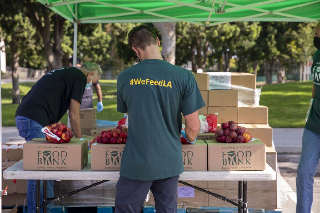 A volunteer in a #WeFeedLA shirt sorts fresh produce at a Los Angeles Regional Food Bank outdoor station. Boxes labeled 