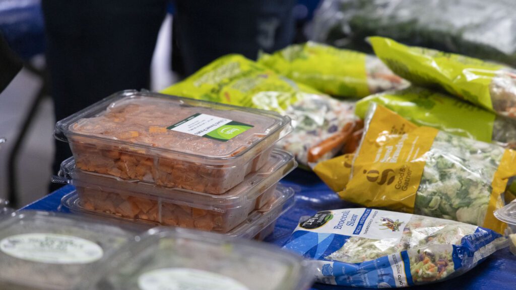 A table with various packaged foods from the Los Angeles Regional Food Bank, including pre-cut vegetables and salad mixes. Some packages are stacked and labeled, displayed against a blue cloth backdrop.
