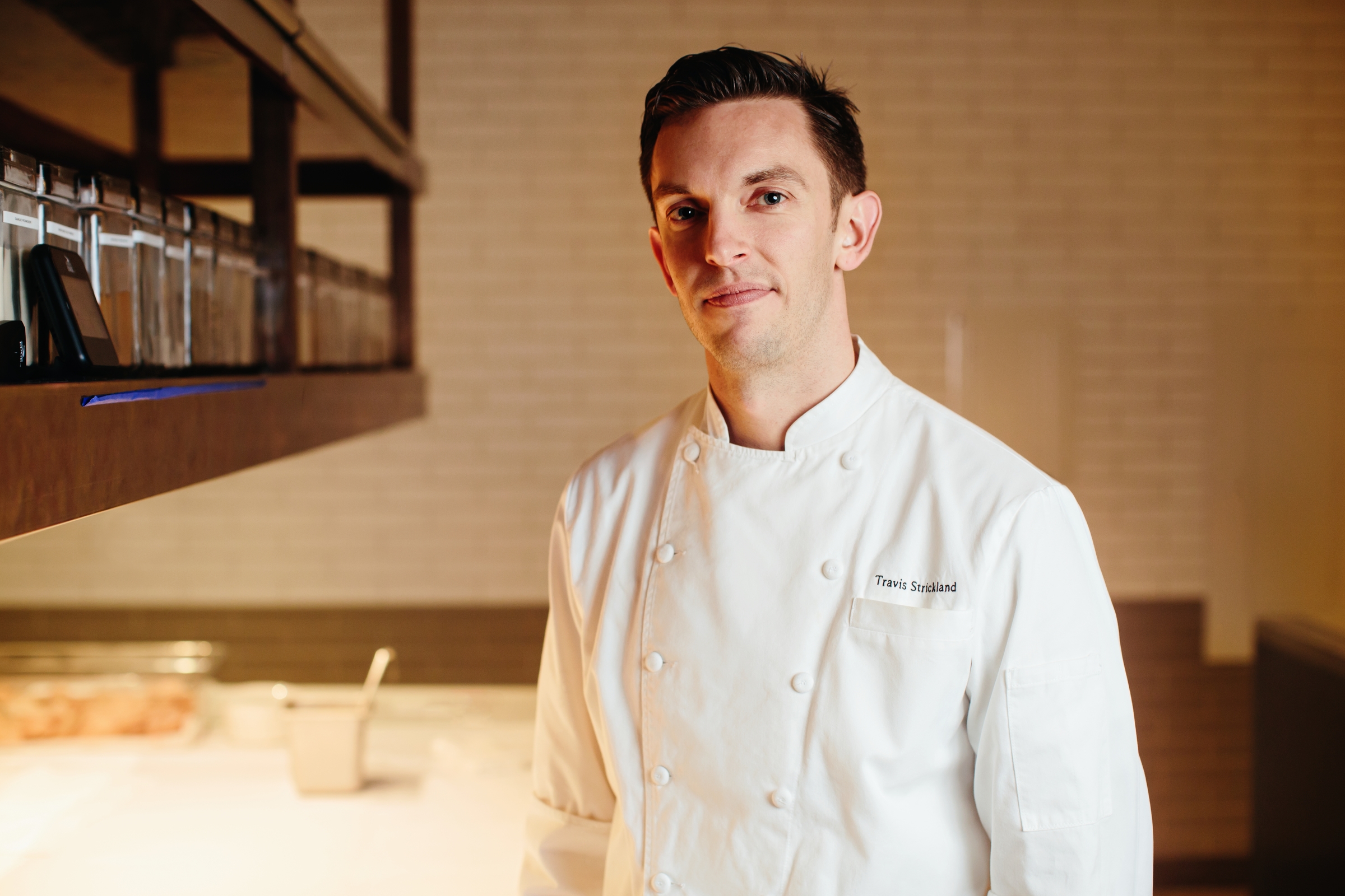 A chef in a white uniform stands in a kitchen with a calm expression, representing the heart of the Los Angeles Regional Food Bank. The background shows shelves with containers and a brightly lit workspace, bustling with purpose and community spirit.