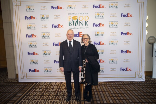 A man and woman stand smiling in front of a step-and-repeat banner with logos for Los Angeles Regional Food Bank, FedEx, and Susan Shelving. They are dressed in formal attire, and the carpet has a geometric pattern.