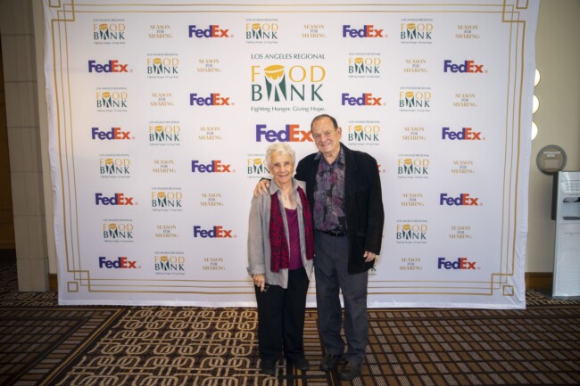 An elderly couple stands smiling in front of a backdrop featuring logos for the LA Regional Food Bank, FedEx, and Susan G. Komen. The man and woman are dressed formally, with the woman wearing a red scarf.