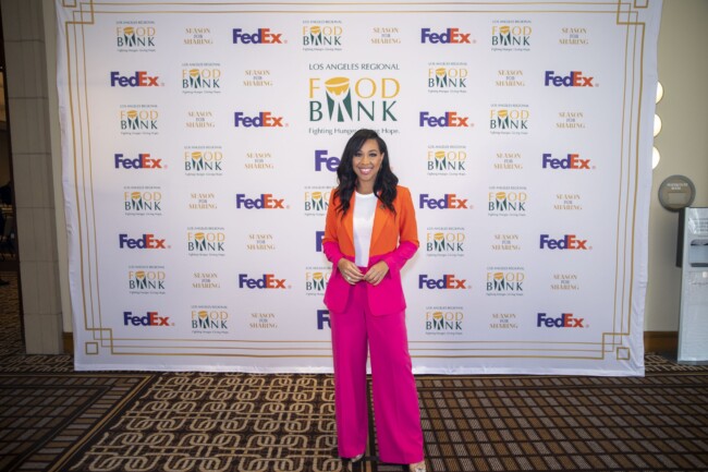 A woman in a vibrant pink suit and orange top stands smiling in front of a backdrop featuring the logos of the Los Angeles Regional Food Bank, FedEx, and Susan Mulling.