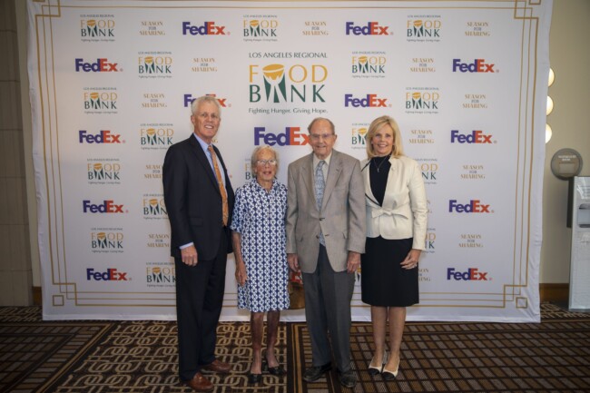 Four people standing in front of a backdrop with logos for the Los Angeles Regional Food Bank, FedEx, and Season of Sharing. They are dressed in formal attire, smiling at the camera on a patterned carpet.