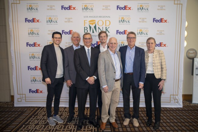 A group of smiling people stands in front of a step-and-repeat banner featuring the logos of the Los Angeles Regional Food Bank and FedEx. Most are wearing business attire, and they pose together on a patterned carpet.