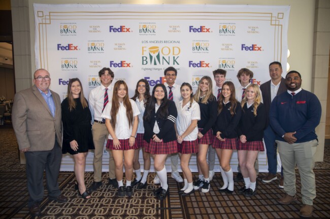 A group of people, including students in plaid uniforms and adults in business attire, stand smiling in front of a Los Angeles Regional Food Bank banner. Logos for FedEx and Susan Maleng are also visible on the banner.