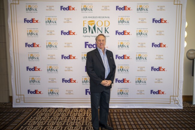 A man in a suit stands smiling in front of a step and repeat banner featuring logos for the LA Regional Food Bank, FedEx, and Season for Sharing. The event appears to be related to charity or community support.