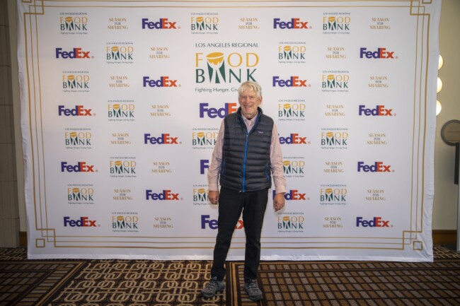 A person standing in front of a backdrop featuring logos of the Los Angeles Regional Food Bank and FedEx. They are wearing a blue puffer vest over a white shirt and black pants, smiling at the camera.