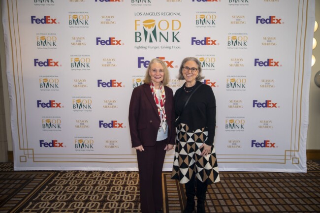 Two women stand smiling in front of a step and repeat banner for the Los Angeles Regional Food Bank, featuring logos from FedEx and Bank of America. They are dressed in business attire.