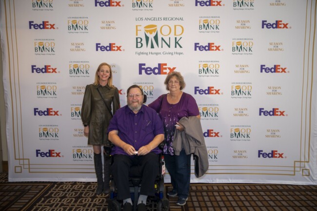 Three people pose in front of a Los Angeles Regional Food Bank backdrop. Two women stand on either side of a man seated in a wheelchair. The background features logos of the food bank and FedEx.