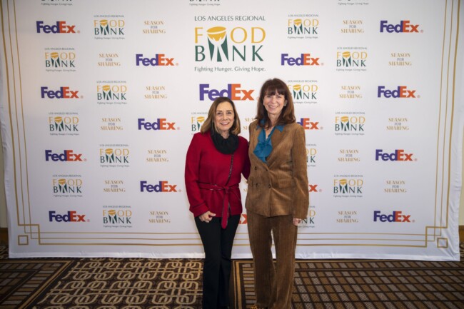 Two people stand smiling in front of a backdrop featuring logos for the Los Angeles Regional Food Bank, FedEx, and Season of Sharing. They are dressed in formal attire, including a red jacket and a brown suit.